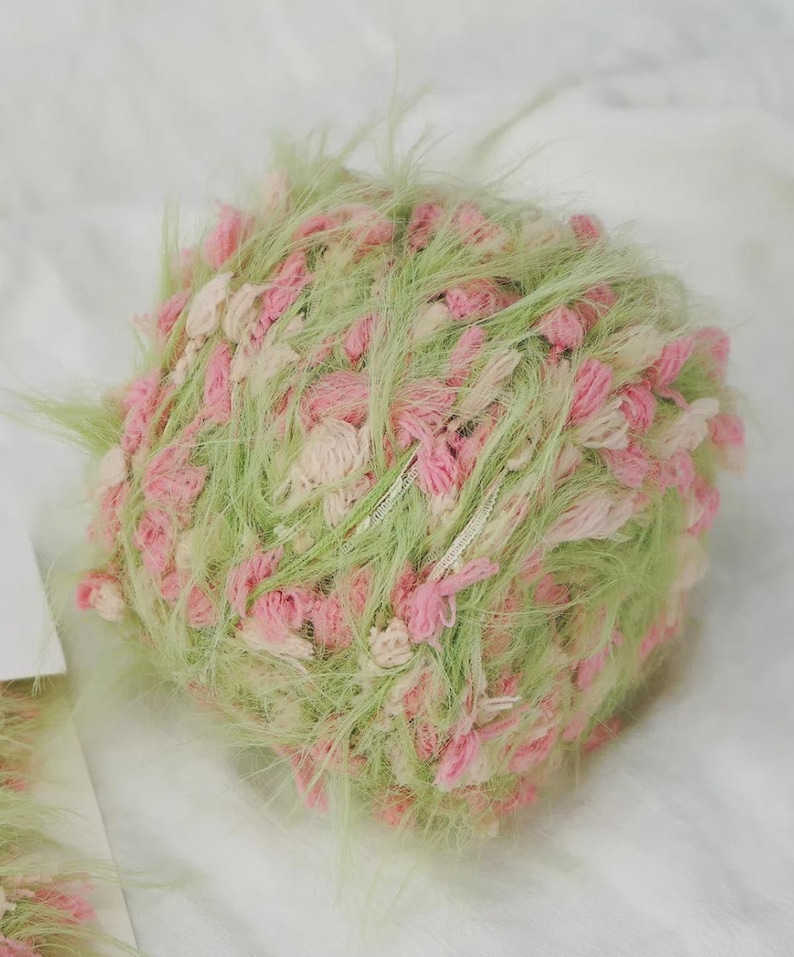 Pode incluir: Novel de fio texturizado em tons pastel de verde, rosa e creme. O fio tem uma apar&ecirc;ncia fofa e difusa com tufos de rosa e creme. Adequado para projetos de tric&ocirc; ou croch&ecirc;, adicionando textura e cor.
