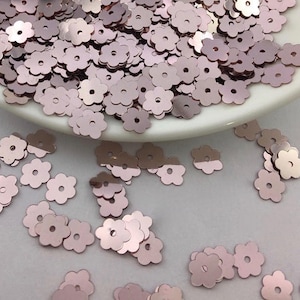 May include: Rose gold flower shaped sequins. The sequins are scattered on a white surface.
