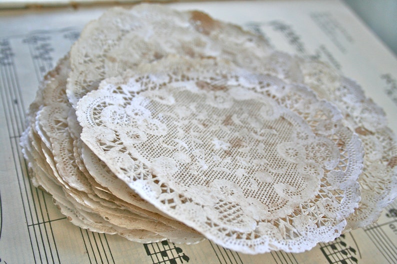 May include: A stack of white paper doilies with a delicate lace pattern. The doilies are arranged in a fan shape and have a slightly aged appearance.