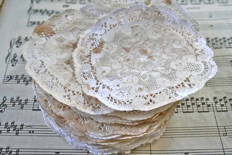 May include: A stack of white lace doilies with a delicate floral pattern. The doilies are slightly brown with age and are arranged on a sheet of music.