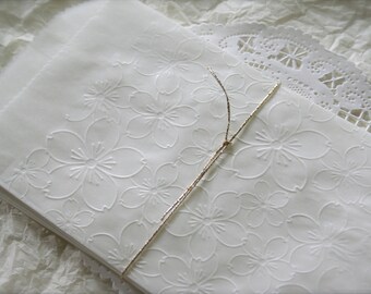 Puede incluir: Bolsa de regalo de papel blanco con un delicado estampado floral en relieve y una cinta dorada.