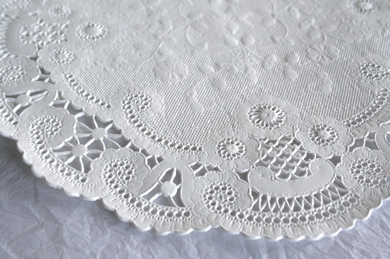K&ouml;nnte beinhalten: Wei&szlig;e Papier-Deko-Serviette mit zartem Spitzenmuster. Die Serviette hat einen gewellten Rand und ein Blumenmuster.