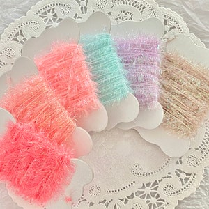 May include: Five spools of iridescent fuzzy yarn in pink, light blue, lavender, beige, and peach colors. The spools are arranged in a fan shape on a white doily.