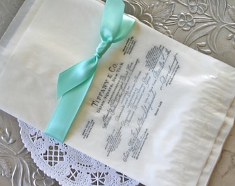 May include: A white paper bag with a light blue satin ribbon tied around it. The bag has a printed label with the text "Tiffany & Co. Union Square, New York" and other details.