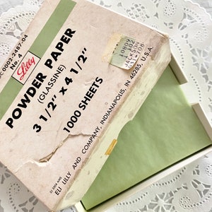 May include: A box of Lilly Powder Paper, 3 1/2 inches by 4 1/2 inches, containing 1000 sheets of glassine paper. The box is white with green and black text. The box is open and the green paper is visible inside.