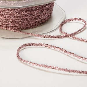 May include: A spool of rose gold glitter trim with a length of trim extending from the spool. The trim is a thin, sparkly, metallic thread.
