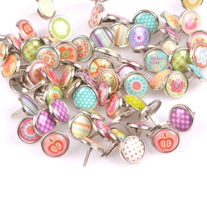 May include: Assortment of colorful patterned metal brads with a silver finish. The brads feature various designs including polka dots, stripes, flowers, and apples. Some brads have the text "miss you" printed on them.