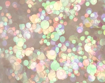 May include: A close-up of iridescent glitter in various shades of pink, green, and blue. The glitter is scattered on a light brown background.