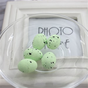 May include: Five small, speckled, green eggs in a clear glass bowl. The eggs have black speckles and are arranged in a cluster.