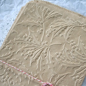 May include: A stack of brown paper gift bags with a holly leaf and berry embossed design. The bags are tied with a red and white twine.