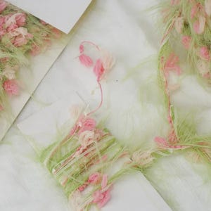 Pode incluir: Close-up de materiais de artesanato decorativos. A imagem apresenta um cart&atilde;o branco com fios rosa e verde e pequenos detalhes de flores rosa. O fio &eacute; enrolado em volta do cart&atilde;o e tem uma textura fofa. O fundo &eacute; branco.