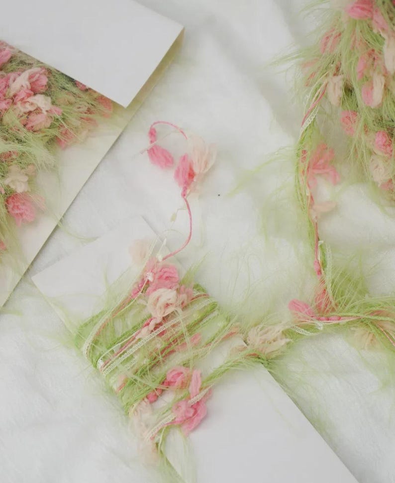 Pode incluir: Close-up de materiais de artesanato decorativos. A imagem apresenta um cart&atilde;o branco com fios rosa e verde e pequenos detalhes de flores rosa. O fio &eacute; enrolado em volta do cart&atilde;o e tem uma textura fofa. O fundo &eacute; branco.