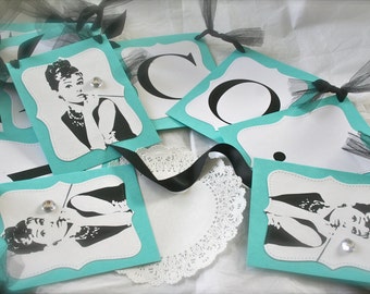 Op de afbeelding: Een turquoise en witte banner met zwart-wit illustraties van Audrey Hepburn. De banner is versierd met zwarte tule en strass. De banner luidt "COCO".