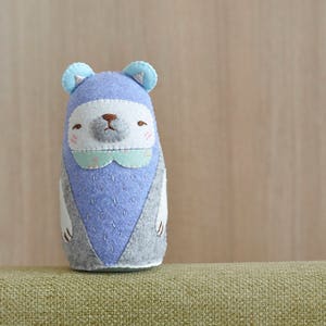 May include: A plush toy shaped like a bear with a blue and gray body, white face, and blue ears. The bear has a light blue collar with a white floral pattern.