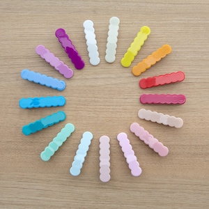 May include: A set of 16 colorful plastic hair clips arranged in a circle on a wooden surface. The clips are all the same size and shape, and they are arranged in a rainbow pattern.