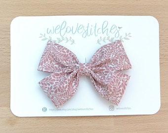 May include: A pink and white patterned fabric bow with a floral design. The bow is on a white card with the text "welovstitches" and a website address.