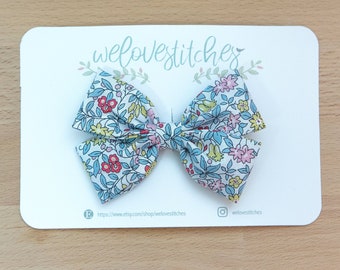 May include: A blue and white floral patterned hair bow with a white background and pink, yellow, and red flowers. The bow is on a white card with the text "welovstitches" and a website address.