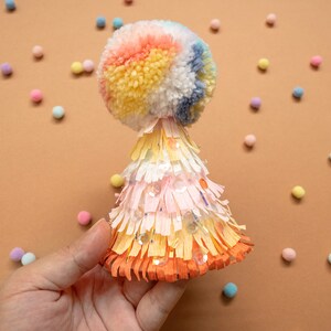 Puede incluir: Un gorro de fiesta colorido con un pompón en la parte superior. El gorro está hecho de papel de seda y tiene una franja alrededor de la parte inferior. El pompón está hecho de hilo y es multicolor.