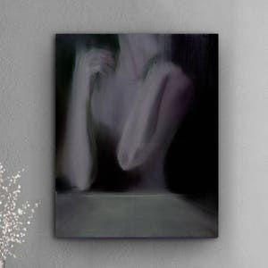 May include: An abstract painting of a figure with arms crossed, in shades of gray and black, hangs above a white cabinet with gold legs. A clear vase with white branches sits on the cabinet, next to a stack of books.