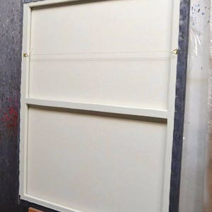 May include: Back of a framed canvas painting, mounted on a wooden easel. The canvas is white, with a dark gray frame. The back of the canvas has a wooden support structure and hanging hardware.