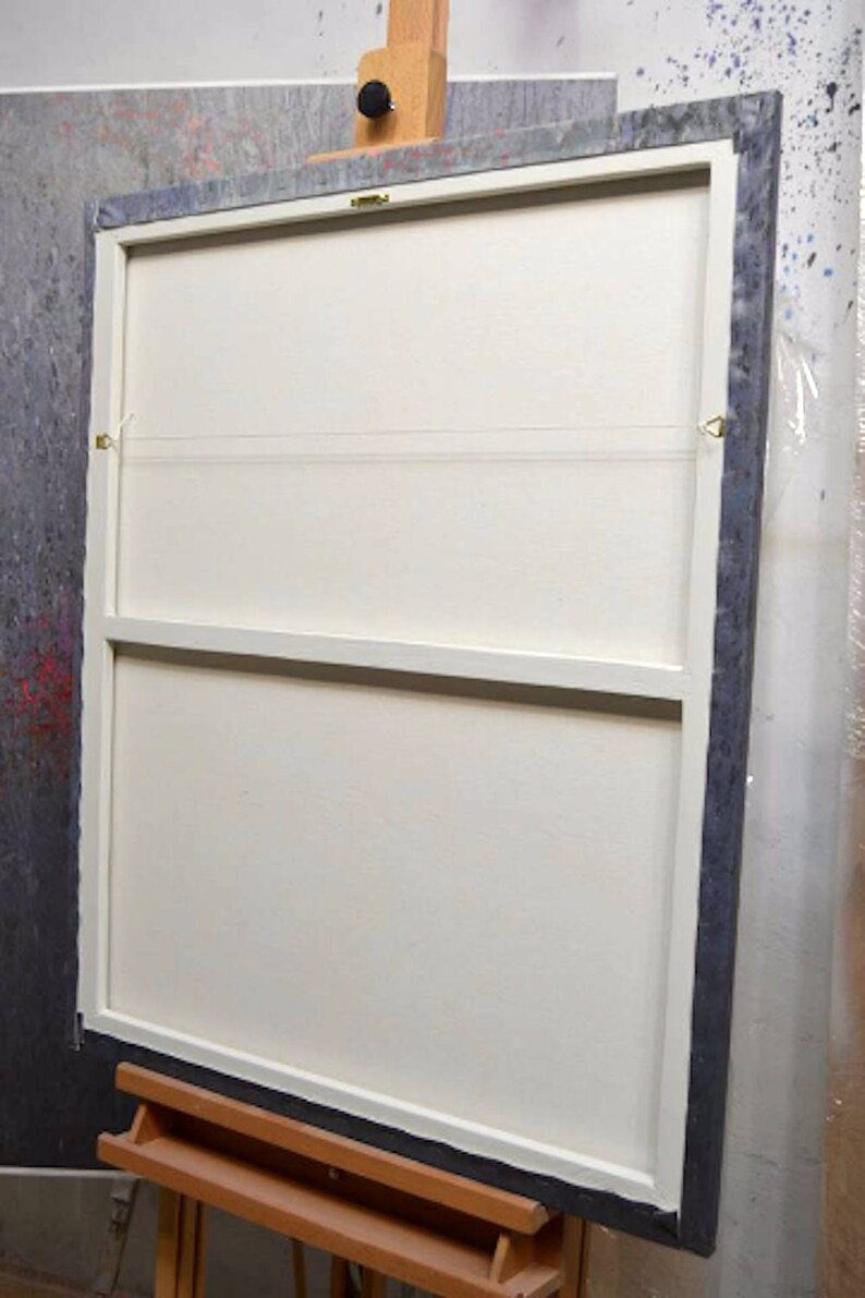May include: Back of a framed canvas painting, mounted on a wooden easel. The canvas is white, with a dark gray frame. The back of the canvas has a wooden support structure and hanging hardware.