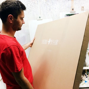 May include: A man in a red t-shirt holds a large, rectangular cardboard package. The package has the text "YURI PYSAR" printed in white. The background includes an easel and art supplies.