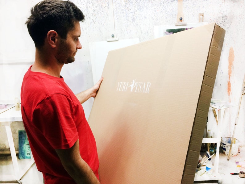 May include: A man in a red t-shirt holds a large, rectangular cardboard package. The package has the text "YURI PYSAR" printed in white. The background includes an easel and art supplies.