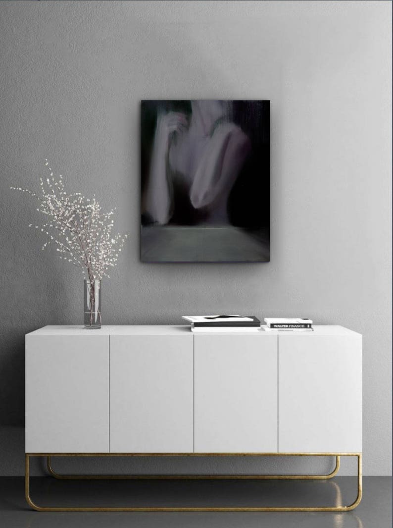 May include: An abstract painting of a figure with arms crossed, in shades of gray and black, hangs above a white cabinet with gold legs. A clear vase with white branches sits on the cabinet, next to a stack of books.