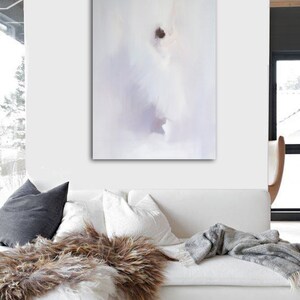 May include: Abstract art piece in a white room. The artwork features soft, blurred shapes in white, gray, and purple hues. A white sofa with pillows and a faux fur throw is in front of the painting. A gray blanket is draped over the sofa.