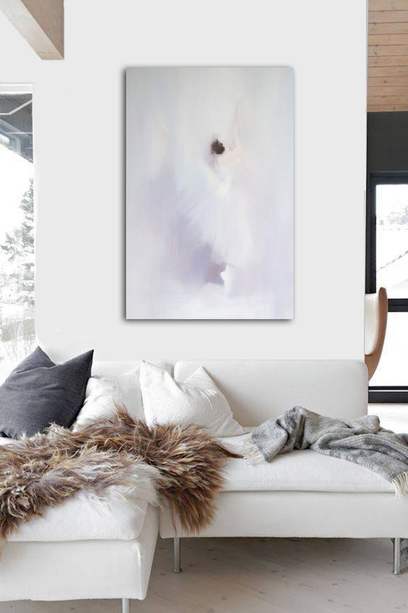 May include: Abstract art piece in a white room. The artwork features soft, blurred shapes in white, gray, and purple hues. A white sofa with pillows and a faux fur throw is in front of the painting. A gray blanket is draped over the sofa.