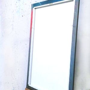 May include: A wooden artist's easel with a blank white canvas. The canvas is framed with a dark blue border. The easel is made of light brown wood and is positioned against a white wall with paint splatters.