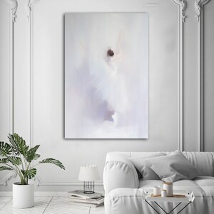 May include: An abstract painting in soft white, lavender, and brown tones hangs on a white wall. The artwork depicts a blurred figure, possibly a dancer, with a flowing white dress. The room features a white sofa, a potted plant, and a small table with decorative items.