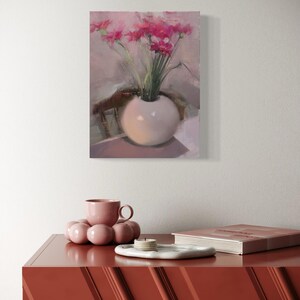 May include: A still life painting of pink flowers in a white spherical vase. The painting hangs above a red-orange cabinet. On the cabinet are a pink teacup, a decorative pink sphere, a white tray with candles, and a book.
