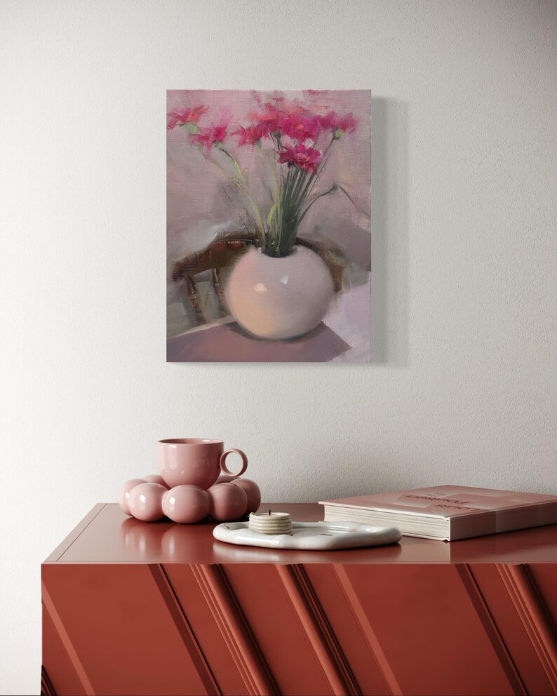 May include: A still life painting of pink flowers in a white spherical vase. The painting hangs above a red-orange cabinet. On the cabinet are a pink teacup, a decorative pink sphere, a white tray with candles, and a book.