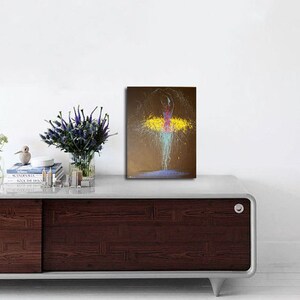 May include: An abstract painting of a ballerina in motion. The figure is depicted with a pink bodice and a yellow tutu, with splashes of blue and white paint creating the illusion of movement. The background is a muted brown.