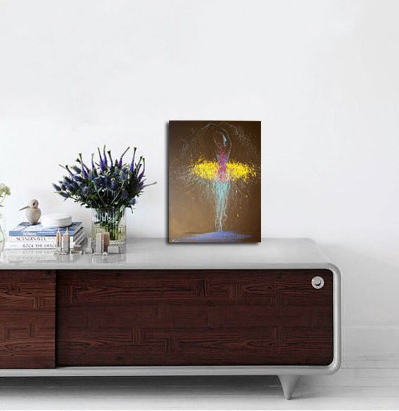 May include: An abstract painting of a ballerina in motion. The figure is depicted with a pink bodice and a yellow tutu, with splashes of blue and white paint creating the illusion of movement. The background is a muted brown.