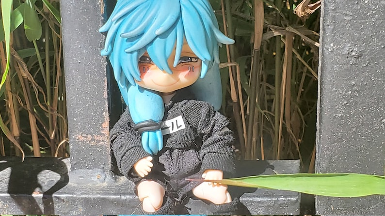 Op de afbeelding: Een kleine pop met blauw haar die een zwart hoodie draagt met het woord "九" op de voorkant. De pop zit op een zwart metalen hek met een groen blad ervoor.