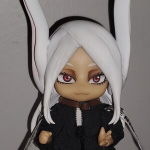 Op de afbeelding: Een wit en zwart anime-figuur met lang wit haar, draagt ​​een zwart jack met een rits en witte bies, en een wit konijnenoren-hoofdband. Het figuur heeft rode ogen en een bruin leren halsband.