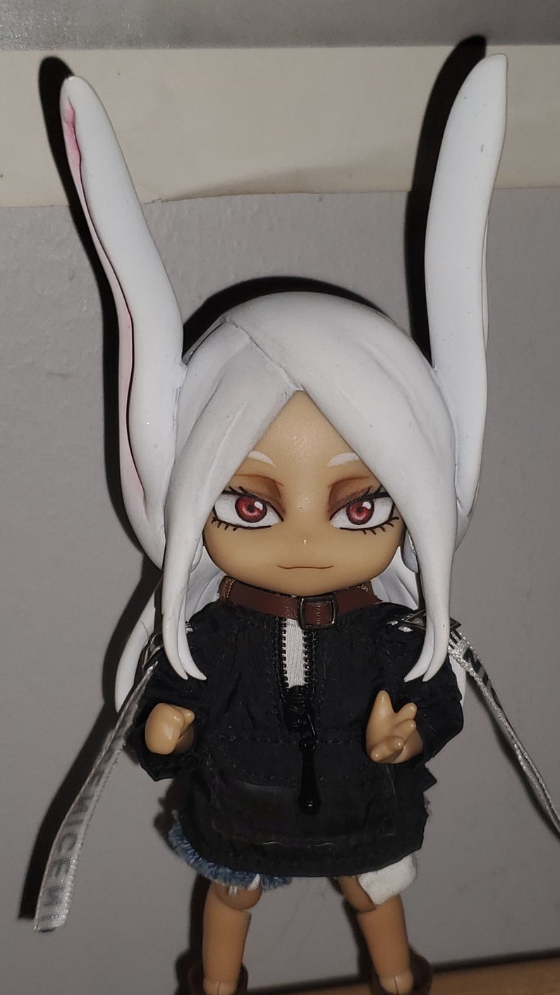 Op de afbeelding: Een wit en zwart anime-figuur met lang wit haar, draagt ​​een zwart jack met een rits en witte bies, en een wit konijnenoren-hoofdband. Het figuur heeft rode ogen en een bruin leren halsband.
