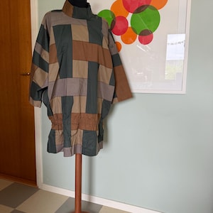 Op de afbeelding: Een tuniek met lange mouwen en een geometrisch blokpatroon in tinten donkergroen, bruin en grijs. Het kledingstuk heeft een hoge kraag, een geplooide elastische taille en een losse, vierkante pasvorm, gepresenteerd op een houten standaard.