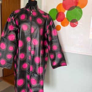 May include: A black raincoat with a vibrant pink polka dot pattern and a full-length zipper. The coat is displayed on a wooden mannequin, showcasing its long sleeves and high collar. The background features a colorful abstract art piece.