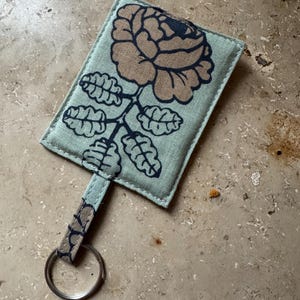 May include: A rectangular key holder with a floral design. The key holder is light green with a large brown and navy blue rose illustration. It has a matching strap and a silver key ring. The key holder is made of fabric.