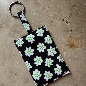 May include: A black and white floral print keychain with a silver key ring. The fabric features white flower shapes with green centers. The keychain has a rectangular pouch shape with a matching fabric loop.