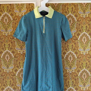 May include: A teal and white striped short-sleeved dress with a yellow collar and a zip closure. The dress is hanging on a white hanger against a patterned background.