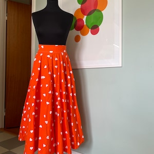 Può includere: Una gonna arancione brillante con un motivo a cuori bianchi. La gonna è ampia e fluida, ed è esposta su un manichino.