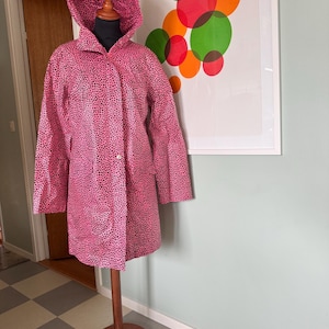 May include: A pink raincoat with a black polka dot pattern and a hood. The raincoat is on a wooden mannequin stand.