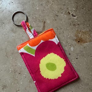 May include: A rectangular fabric keychain with a floral pattern in shades of pink, orange, green, and yellow. The keychain has a silver metal ring attached to a fabric loop. The fabric is stitched with white thread.