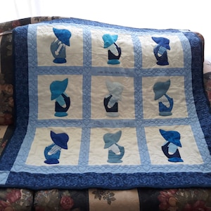 May include: A blue and white quilt with a pattern of nine embroidered figures wearing blue bonnets. The quilt is stitched with a heart pattern and has a blue border.