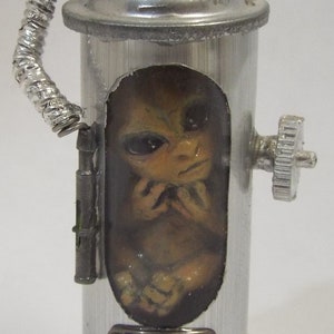 May include: A silver metal container with a small, brown alien figure inside. The container has a hose attached to the top and a knob on the side. There are three colored buttons on the front of the container.