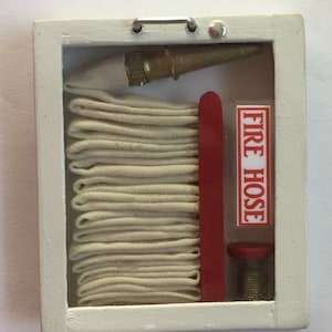 May include: A white wooden frame with a glass front containing a rolled up white fire hose, a red fire hose nozzle, and a brass fire hose connector. The frame has a red label that says "FIRE HOSE".
